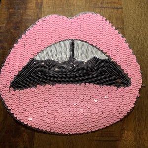 May include: Pink sequined lips with an open mouth design. The lips are outlined in pink sequins, with a black sequined interior and a silver sequined tongue. The item is likely a decorative pillow or accessory.