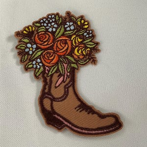 May include: Embroidered patch featuring a brown cowboy boot filled with a bouquet of flowers. The flowers include orange roses, yellow blooms, and small blue and white blossoms, all with green leaves. The boot has a pink trim.