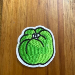 May include: Embroidered patch featuring a green bell pepper design with a white border and black outline. The patch is on a wooden surface. The patch is a vibrant green color.