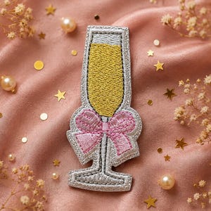 May include: An embroidered patch shaped like a champagne flute with a yellow liquid fill, a silver stem and base, and a pink bow tied around the middle. The patch features detailed stitching with a silver border and a textured finish throughout.