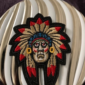 May include: Embroidered patch featuring a Native American chief with a headdress. The headdress is made of red, yellow, and white feathers. The chief's face is brown with red and yellow accents. The patch is black with a white background.