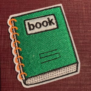 May include: Embroidered green notebook patch with the word "book" in a white rectangle. The patch has a white border and orange spiral binding. The notebook design is a fun accessory for bags, jackets, or other fabric items.