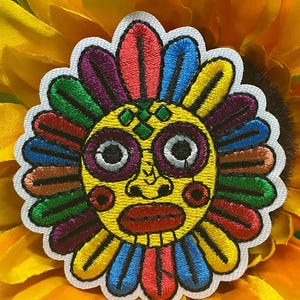 May include: Embroidered patch featuring a colorful sun face design with a yellow background, surrounded by a sunflower. The sun face has large eyes, a red mouth, and colorful rays in shades of red, blue, green, and purple.
