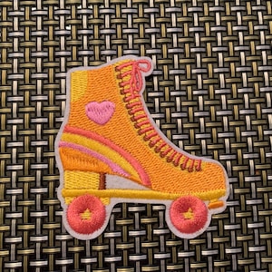 May include: An embroidered patch shaped like a high-top roller skate. The skate is primarily yellow and orange with pink laces, a pink heart detail on the side, and two pink wheels. The patch has a white border and a textured, stitched appearance.