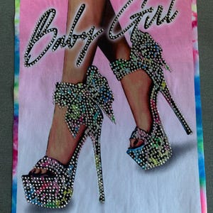 May include: A pink and white tie-dye fabric with the text "Baby Girl" in black rhinestones. Two high-heeled shoes with bows are decorated with colorful rhinestones.