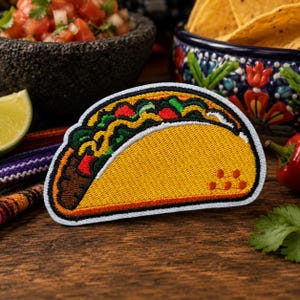 May include: A taco-shaped embroidered patch featuring a golden yellow shell, brown meat, green lettuce, and red tomato toppings. The design includes a black border and a small cluster of orange dots on the lower right side of the taco shell.