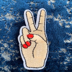 May include: Embroidered peace sign patch with a hand making the peace sign gesture. The hand is beige with red nail polish.
