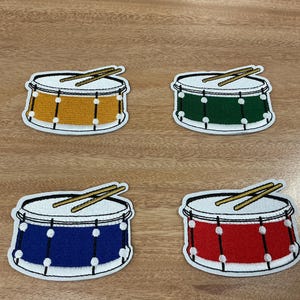 May include: Four embroidered drum patches in yellow, green, blue, and red. Each patch features a white drumhead, a white border, and two drumsticks. The drums have white accents and are set against a wooden background.