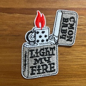 May include: A gray and white embroidered patch of a lighter with the text "Light My Fire" and "Cmon Baldi" on the side. The lighter has a red flame.