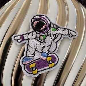 May include: An embroidered iron-on patch of an astronaut wearing a white spacesuit with a purple, pink, and green helmet, skateboarding on a purple skateboard with yellow wheels.