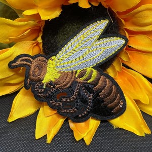 May include: Embroidered bee patch with yellow, brown, and black details, set against a bright yellow sunflower. The bee patch features intricate stitching and a black border, creating a striking contrast.