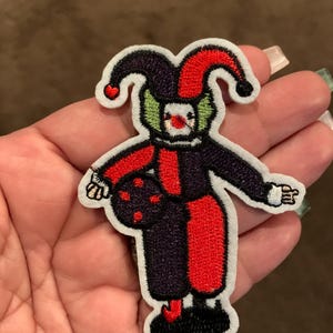May include: An embroidered iron-on patch of a jester with a red and black costume, green face, and red and white hat.
