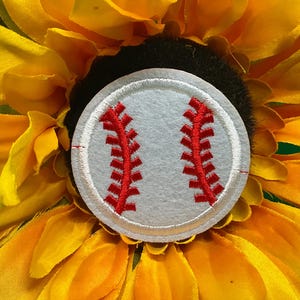 May include: A baseball-themed accessory featuring a white baseball patch with red stitching, set against a backdrop of a vibrant yellow sunflower. The baseball patch is circular and detailed.