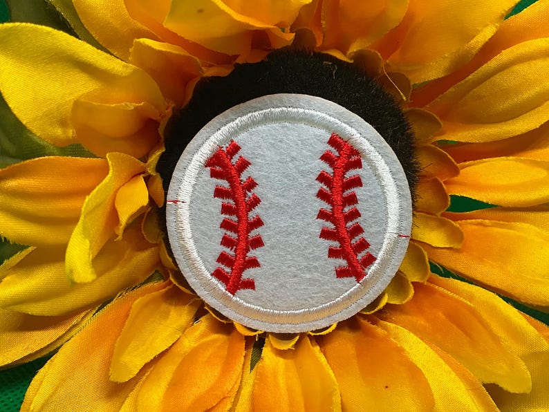 May include: A baseball-themed accessory featuring a white baseball patch with red stitching, set against a backdrop of a vibrant yellow sunflower. The baseball patch is circular and detailed.