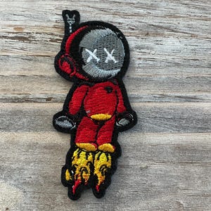 May include: Embroidered patch featuring a cartoon astronaut in a red spacesuit with a gray helmet and crossed-out eyes. The astronaut has yellow flames coming from the bottom of the boots. The patch is outlined in black.