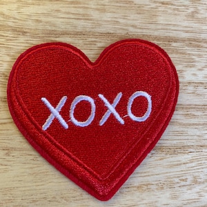 May include: A red heart-shaped patch with the embroidered text "XOXO" in white. The patch is made of a textured fabric and has a stitched border. The background is a light-colored wooden surface.