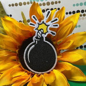 May include: A black and white embroidered patch of a cartoon bomb with a yellow explosion design. The patch is placed on a bright yellow sunflower with green leaves. The background is a patterned surface.
