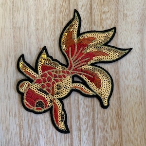 May include: Embroidered patch in the shape of a goldfish. The patch features red and gold sequins, with black outlining. The fish has a detailed design, with a black border.