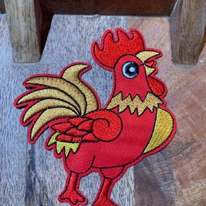 May include: Red embroidered rooster patch with a gold tail and a white eye. The rooster is standing on a wooden surface.