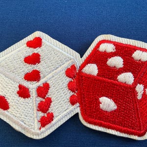 May include: Two embroidered dice patches. One die is white with red heart shapes, the other is red with white dots. Both have a white border and are set against a blue background.