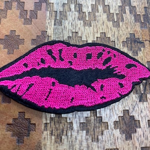 May include: Embroidered patch in the shape of a pair of lips. The lips are bright pink with black outlining. The patch is on a wooden surface with a geometric pattern.