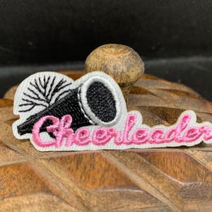 May include: Embroidered patch featuring a black megaphone and white pom-pom design. The word "Cheerleader" is embroidered in pink script. The patch is on a white background.