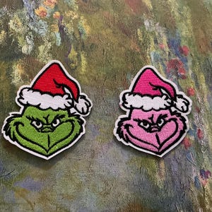 May include: Two embroidered patches featuring a cartoon character's face wearing a Santa hat. One patch is green with a red hat and white trim, the other is pink with a pink hat and white trim. Both have black outlines.