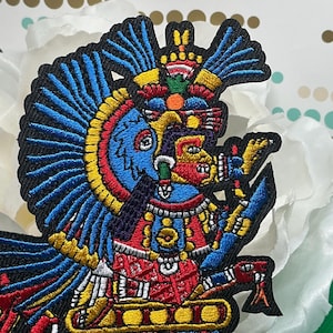 May include: An embroidered patch featuring a colorful depiction of an Aztec deity with a large blue feathered headdress, red and yellow accents, and intricate patterns. The figure is shown in profile with a serpent-like creature at the base.