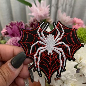 May include: Embroidered patch featuring a white spider on a black web with red accents. The spider patch is in the shape of a spider web. The patch is held in front of a bouquet of flowers.