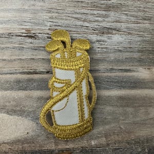 May include: Embroidered patch featuring a golf bag with clubs. The bag is white with gold trim and a gold handle. The golf clubs are gold. The patch is likely for a jacket, bag, or other apparel item.