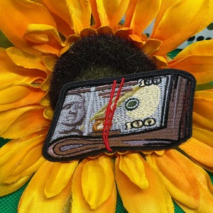 May include: A rectangular embroidered patch featuring a stack of one hundred dollar bills secured with a thin red band. The design includes detailed stitching in shades of grey, brown, and yellow, with the number 100 clearly visible on the currency.