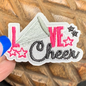 May include: A white embroidered patch with the words "LOVE Cheer" in pink and black. The patch features a megaphone, stars, and a decorative font, perfect for cheerleading enthusiasts.