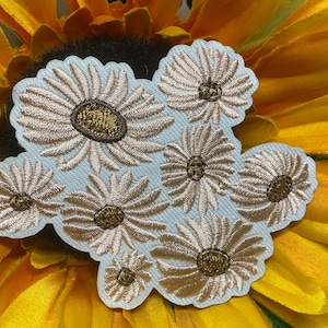 May include: An embroidered patch featuring a cluster of eight daisy flowers with light beige petals and dark gold centers. The flowers are arranged on a light blue fabric backing with a scalloped edge that follows the outline of the floral design.