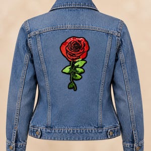 May include: A blue denim jacket featuring a large, centered rose applique on the back. The rose is constructed from red and green sequins, depicting a detailed flower head with a stem and leaves. The jacket has a classic collar and button cuffs.