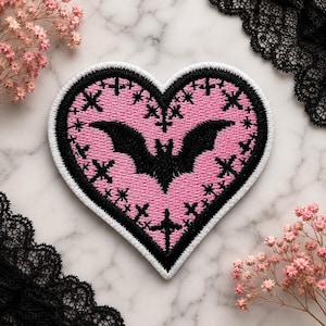 May include: A heart-shaped embroidered patch featuring a pink center with a black bat silhouette at the middle. The design includes small black cross motifs scattered across the pink background, all enclosed by a thick black border and a white edge.
