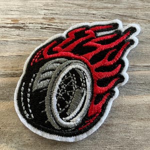 May include: Embroidered patch featuring a black tire with a silver rim, surrounded by red flames. The patch has a white border. This patch is suitable for jackets, bags, or other fabric items.