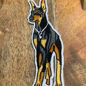 May include: Embroidered patch of a black and tan Doberman Pinscher with a diamond necklace. The dog is standing with its front legs crossed and its head tilted to the side.