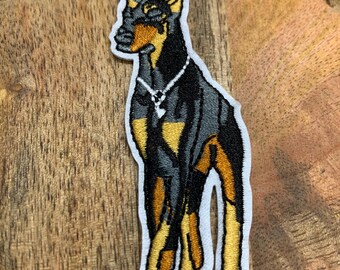 May include: Embroidered patch of a black and tan Doberman Pinscher with a diamond necklace. The dog is standing with its front legs crossed and its head tilted to the side.