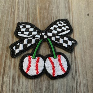 May include: Embroidered patch featuring a bow with a black and white checkered pattern, above two cherries. The cherries are white with red stitching, green stems, and a black border. The patch is on a wood grain surface.