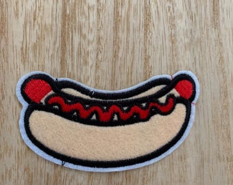 May include: A colorful patch featuring a cartoon hot dog. The hot dog has a light tan bun, red ketchup, and red end pieces. The patch is outlined in black and white.