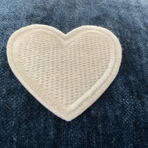 May include: A white heart-shaped patch with a stitched border. The patch is made of a textured fabric and is set against a blue background. This patch could be used for clothing or accessories.
