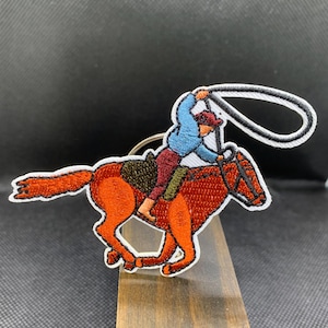 May include: Embroidered patch featuring a cowboy on a running horse, with a lasso in hand. The horse is a vibrant orange, and the cowboy wears a blue shirt and red hat. The patch has a white border.
