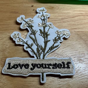 May include: An embroidered patch featuring a white background with a botanical illustration of wildflowers in olive green thread. Below the flowers, a rectangular beige banner contains the black embroidered text "Love yourself".