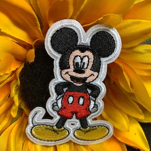 May include: Embroidered patch featuring Mickey Mouse with black ears, a red shorts, and yellow shoes. The patch has a white border and is set against a yellow flower.