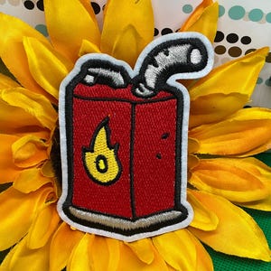 May include: A rectangular red embroidered patch featuring a yellow flame icon on the front, a silver spout at the top, and a thick white border. The design depicts a fuel container with black outlines, showcasing detailed stitching throughout the piece.