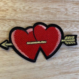 May include: Embroidered patch featuring two red hearts pierced by a gold arrow. The hearts have a pink outline and are bordered by black stitching. The arrow has gold detailing.