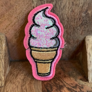 May include: A pink felt ice cream cone patch with a glittery pink and white ice cream swirl. The cone is outlined in black and has a gold texture.