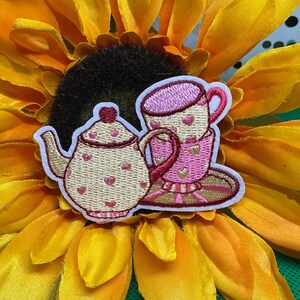 May include: An embroidered patch featuring a cream-colored teapot and a pink teacup with a matching saucer. Both the teapot and the teacup are decorated with small pink heart patterns. The patch has a white border and is placed on a yellow flower.