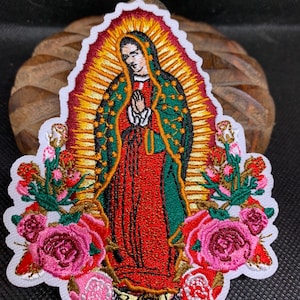 May include: Embroidered patch featuring a religious figure in a red robe and green cloak, surrounded by a golden halo and pink roses. The patch has a white border and is likely for clothing or accessories.
