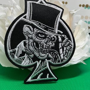 May include: A black and silver embroidered patch shaped like a spade playing card suit. The design features a skeletal figure wearing a top hat, with the skull and hat rendered in detailed silver thread against a solid black background.